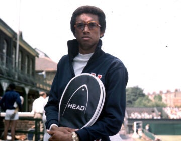 A young Arthur Ashe holding a tennis racket in a cover. He is sitting on the ledge of a balcony at a tennis club.