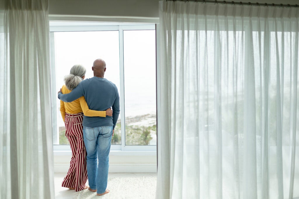 Rear view of active African-american senior couple with arm around standing near window at home