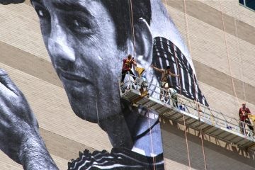 French street artist JR (shirtless) installs his latest mural on the Graham Building in Center City. (Emma Lee/WHYY)