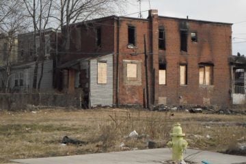 A house with boarded-up windows is visible on an empty lot on a winter's day.