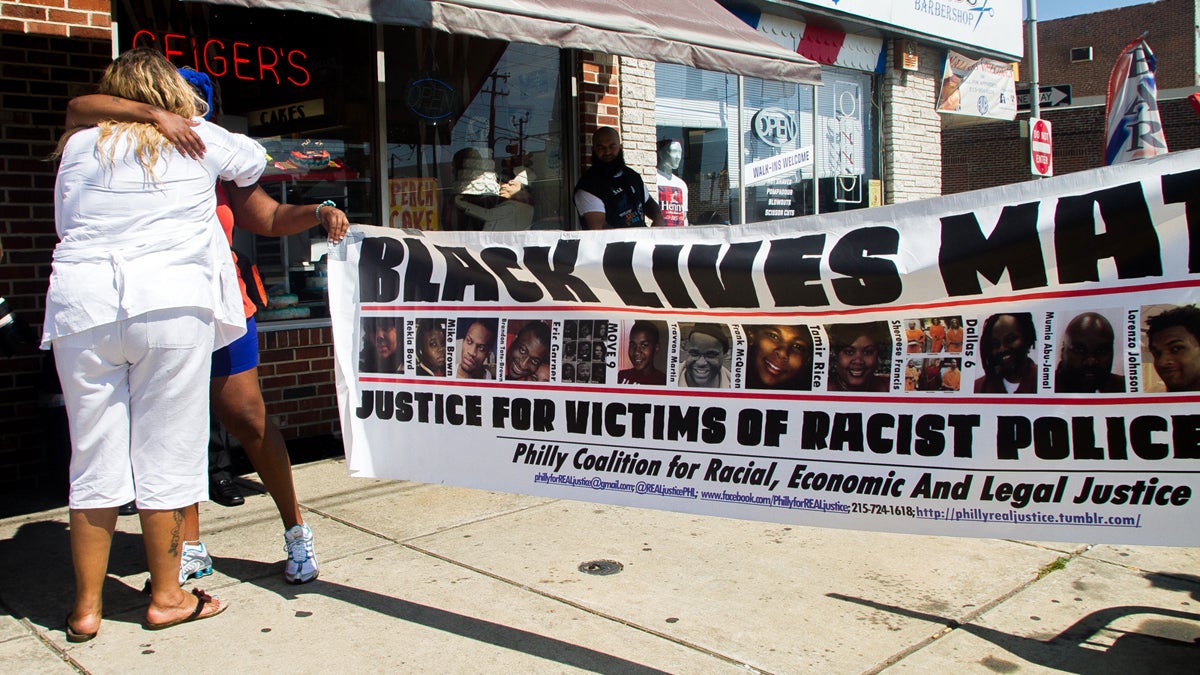 A banner commerating black men and women killed by police near the site where Brandon Tate-Brown was shot by officers in December on Frankford Avenue. Tate-Brown's mother Tanya Brown-Dickerson(in white) is pictured hugging a supporter. (Brad Larrison/for NewsWorks