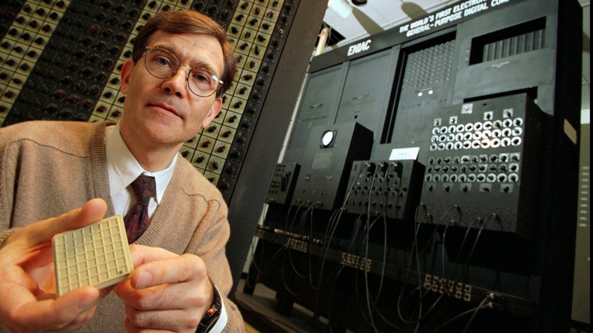 Jan Van der Spiegel holds a microprocessor (the black dot at the lower right corner of the square pad in his hand) in front of about one-tenth of the 50-ton original Electronic Numerical Integrator and Computer (ENIAC)