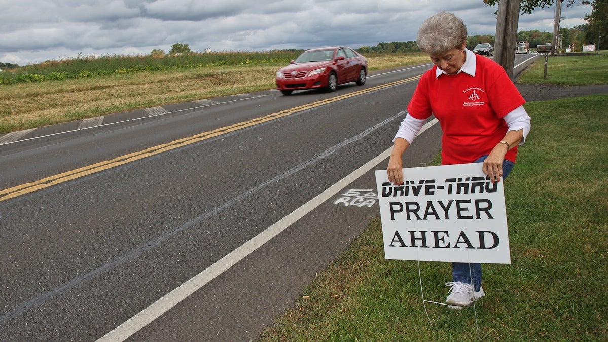 Ruth Portzline is the Pastor of Saint John's United Methodist church in Ivyland, Pa. She stakes a sign inviting motorists to pray with volunteers a few plots down from the church. (Kimberly Paynter/WHYY)