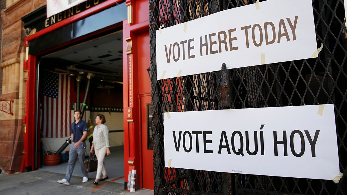 In the background, several people exit a polling station. In the foreground are two signs that read, 