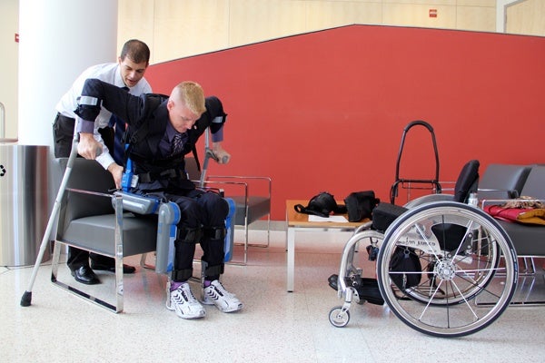 Pierre Asselin assists medical student and Air Force veteran Ian Brown to stand for photos using an exoskeleton that allows paraplegics to walk. (Emma Lee/for NewsWorks)