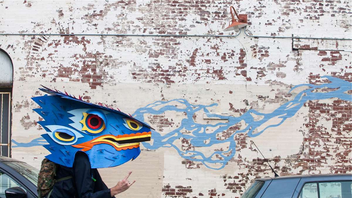 Rob Leef, wearing a dragon mask in the Kinetic Sculpture Derby appears to be breathing the blue flames painted on the brick wall behind him