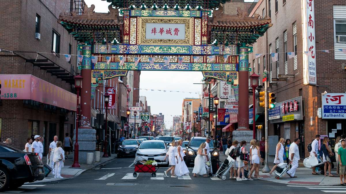 Thousands of people dressed in white, carrying their food and furniture, parade through Chinatown on their way to Dîner en Blanc at Franklin Square Park. (Brad Larrison for NewsWorks)