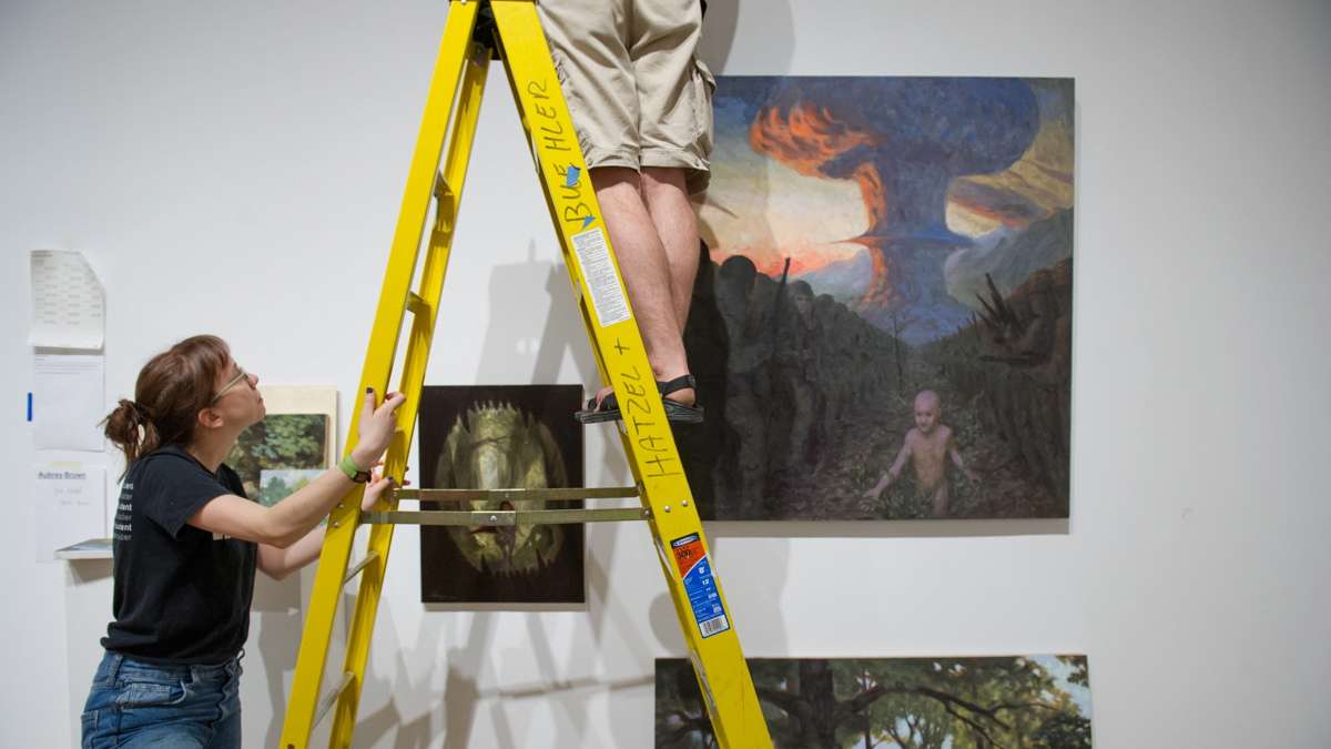 PAFA Alumni Zoe Zurad holds the ladder for Aubrey Brown as he hangs his paintings.