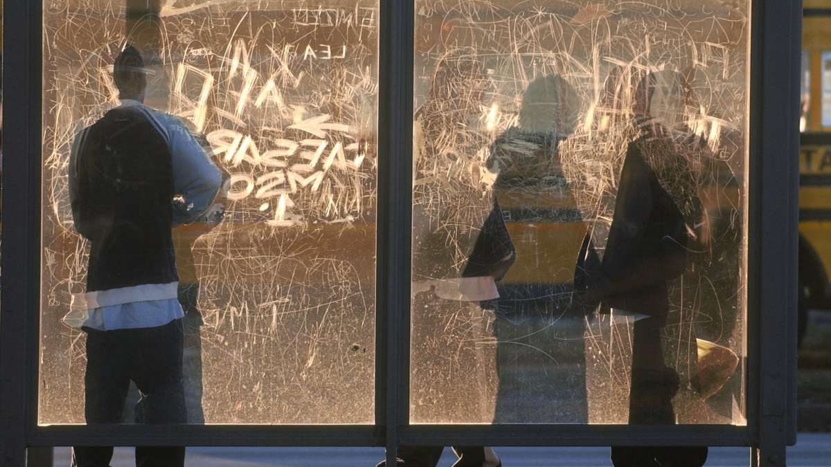 The rising sun throws shadows against the windows of a bus shelter, etched with names and initials.