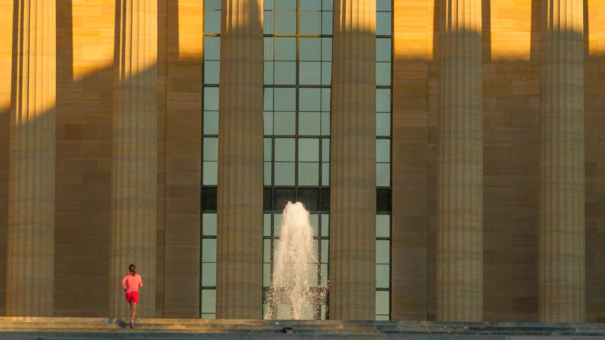 A woman in red shorts and pink shirt runs up the steps of the Philadelphia Art Museum toward the fountain while the rising sun casts long shadows.