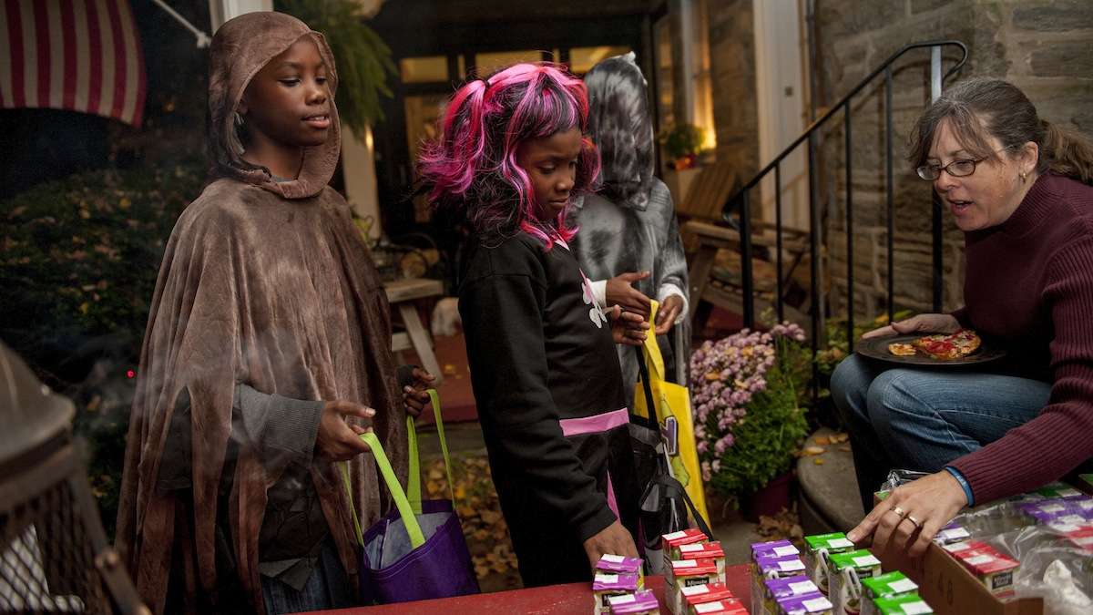 Robin Vallette sits on her porch in Chestnut Hill and gives juice boxes away to trick-or-treaters (from left) Darrius Ward, 11, Daysha Ward, 8, and Taren Williams, 12, all from Chestnut Hill, on Halloween night. (Tracie Van Auken/for NewsWorks)