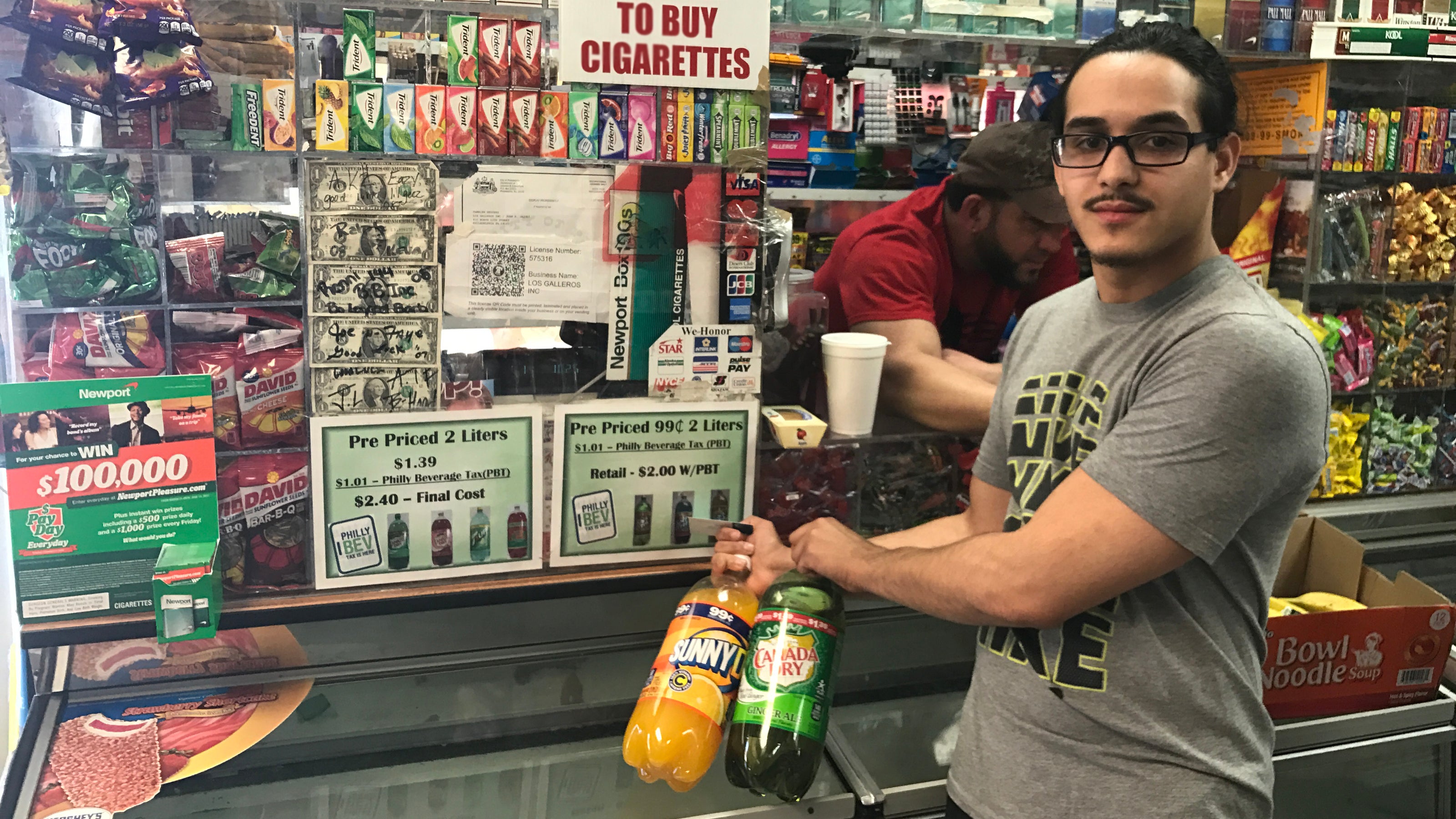  Frayby Perelta holds up 2-liter bottles of soda that are now mislabeled with the wrong price because of Philadelphia soda tax. A sign in the bodega he operates explains that the tax has doubled the price. (Bobby Allyn/WHYY) 