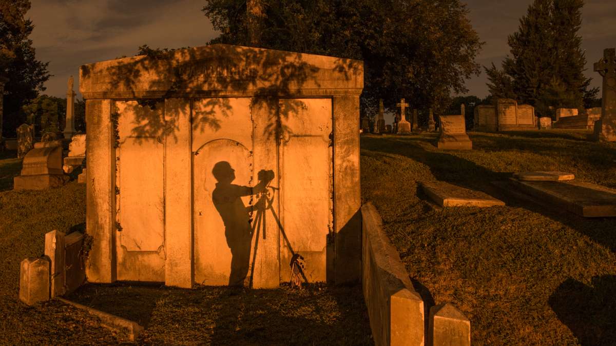 A photographer's shadow is cast against the wall of a mausoleum from the illumination of street lights.