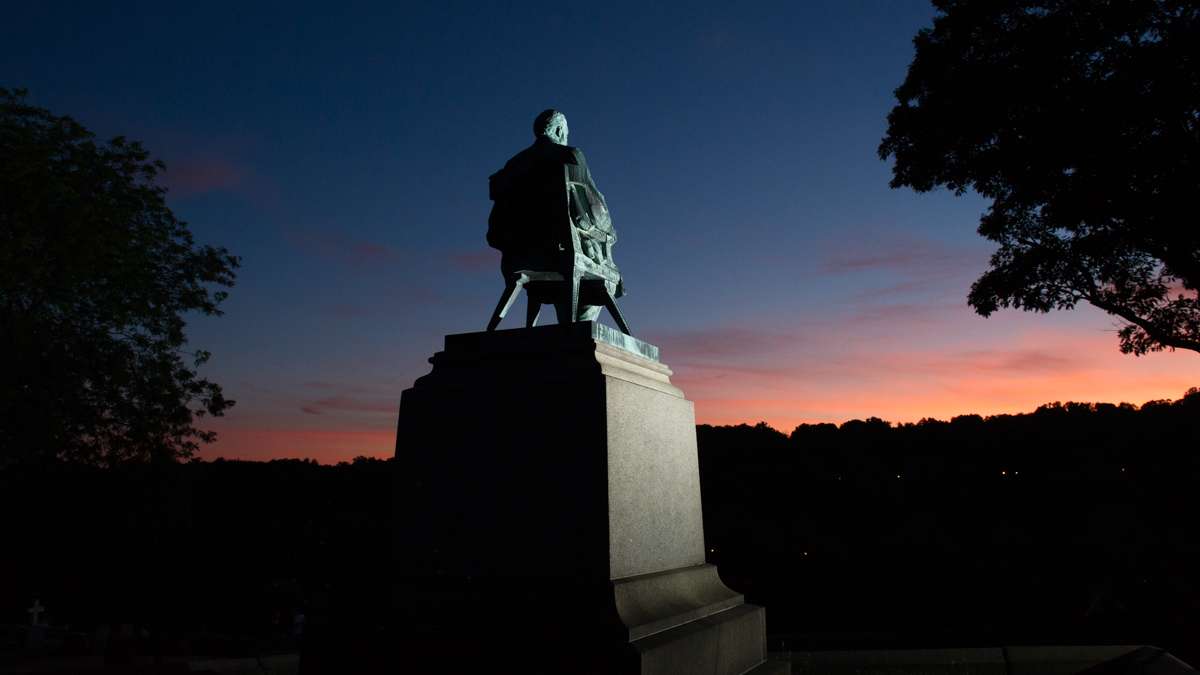 A statue of W. E. Hughes sits atop his burial plot facing the Schuylkill River at sunset.