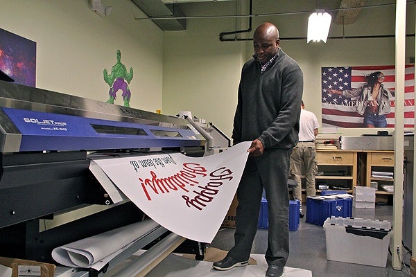 <p>Keith Leaphart, owner of Replica print shop in Center City, watches as a window sign emerges from the printer. (Emma Lee/for NewsWorks)</p>
