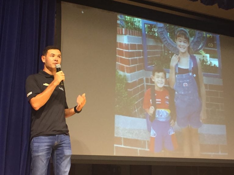 Top NASCAR racer Kyle Larson addresses students Tuesday at t St. Georges Technical High School while a photo with his sister from his youthful racing days is projected on a screen. (Cris Barrish/WHYY)