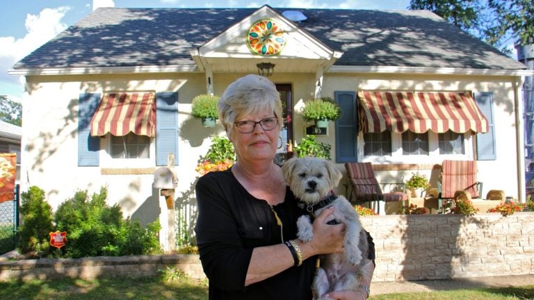 A woman with white hair stands in front of her yellow house, holding her white haired dog