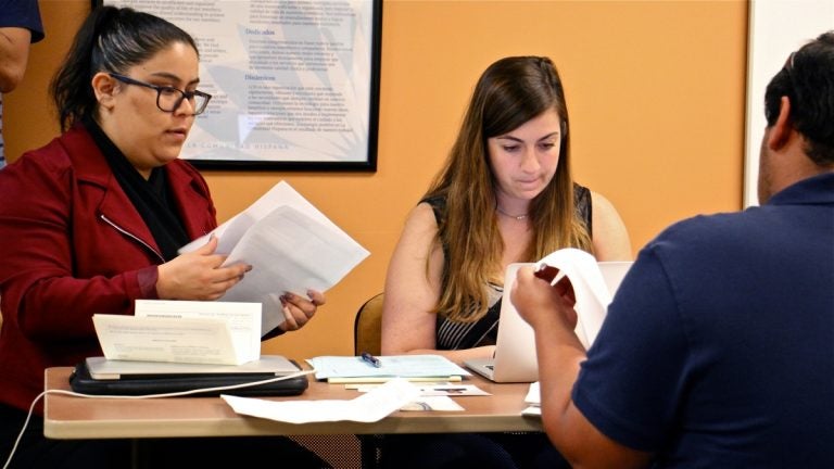 Two women at a desk facing the camera look over paperwork, a man, on the other side of the desk, his back to the camera