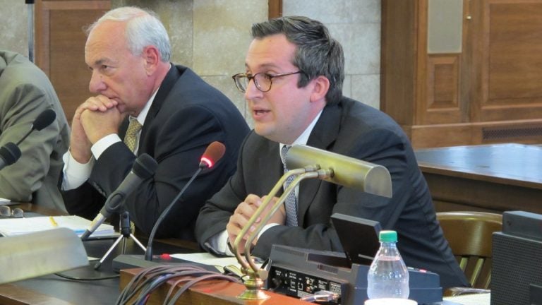 Two men sit at a table, testifying, in front of microphones