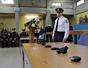 Lt. Tom McLean stands near a table, a half-dozen body camera options are displaued; officers sit and stand in the background