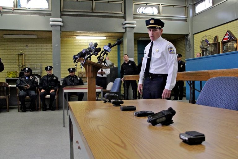 Lt. Tom McLean stands near a table, a half-dozen body camera options are displaued; officers sit and stand in the background