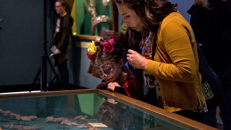 A mother and child look at artifacts in a glass case at Penn Museum. The child's face is painted like a skull.