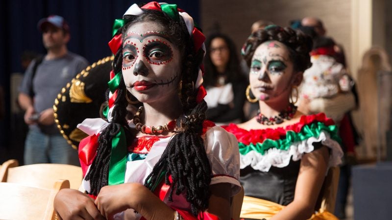 Young girls in red white and green costumes with faces painted like skulls prepare to dance at a Day of the Dead Celebration