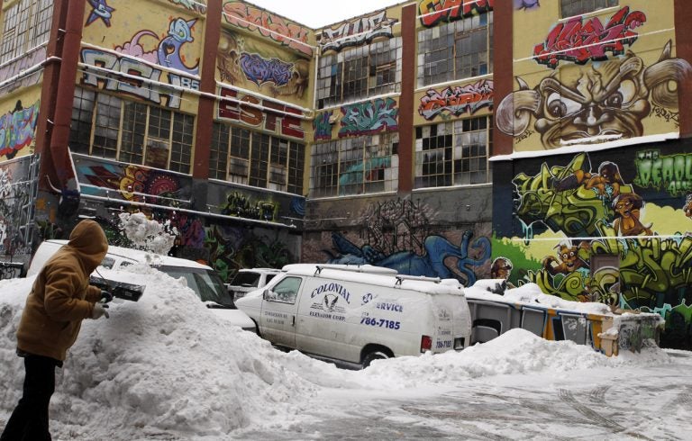 A warehouse covered in large grafiti art; In the foreground, a man shovels snow.
