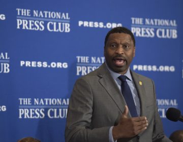 A man in a suit speaks in front of a blue, National Press Club background