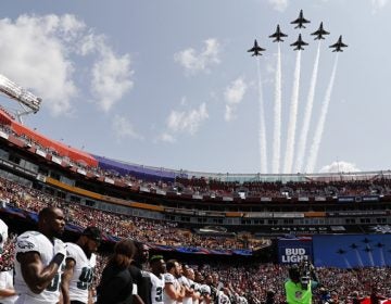 In formation and against a blue sky, members of the U.S. Air Force Thunderbirds fly overhead while Eagles players look on, hands on their hearts, during the national anthem