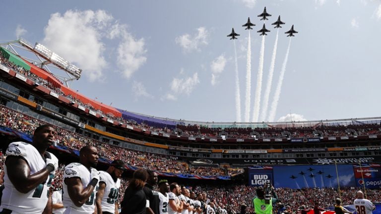 In formation and against a blue sky, members of the U.S. Air Force Thunderbirds fly overhead while Eagles players look on, hands on their hearts, during the national anthem