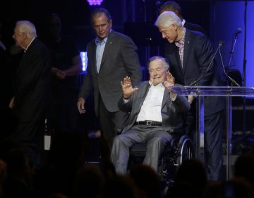 President George H.W. Bush in a wheelchair, hands up, palms out, leaving a stage; President Bill Clinton pushes Bush, Sr.; President George W. Bush walks off stage, looking back to his father