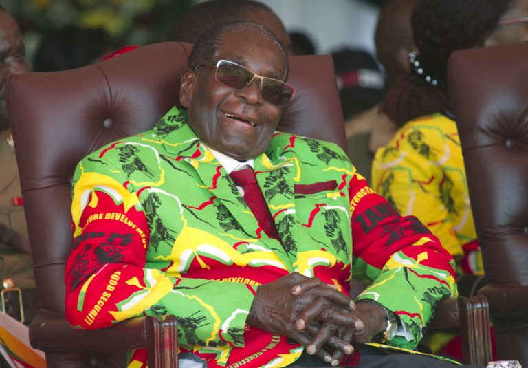 Zimbabwean President Robert Mugabe sits in a chair, sunglasses and a smile, wearing a colorful yellow, red, and green jacket