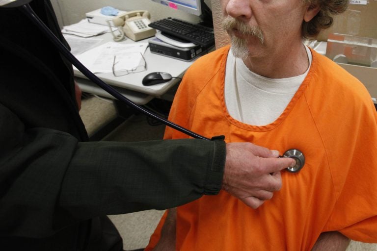 A person holds a stethoscope to the chest of a man in an orange prison jumpsuit