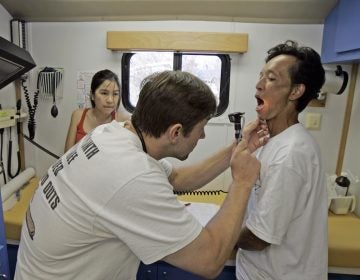 A doctor uses a scope to look into the mouth of a patient; another woman looks on from the rear