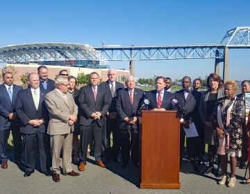 Almost 20 local leaders, politicians, and businesspeople, mainly men in suits stand around a podium; the bacground a bridge and river