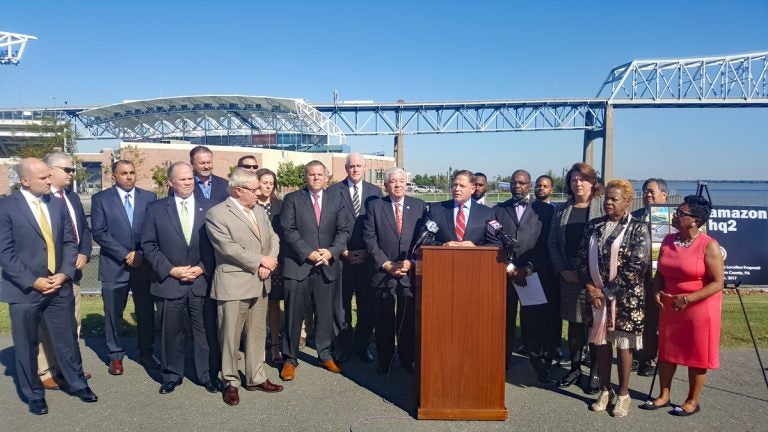 Almost 20 local leaders, politicians, and businesspeople, mainly men in suits stand around a podium; the bacground a bridge and river