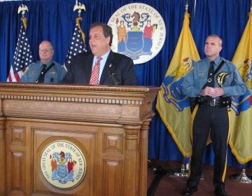 Governer Christie stands at a podium flanked by two officers, American flag, and New Jersey flag, and seal