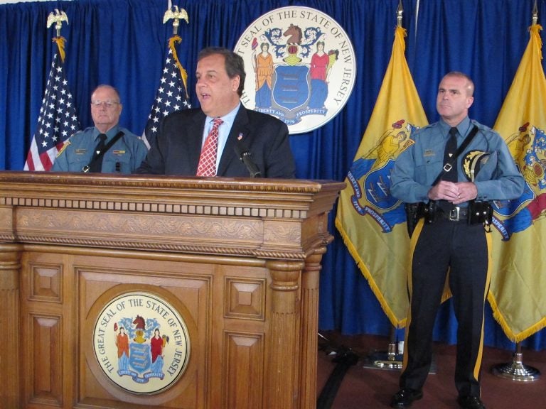 Governer Christie stands at a podium flanked by two officers, American flag, and New Jersey flag, and seal