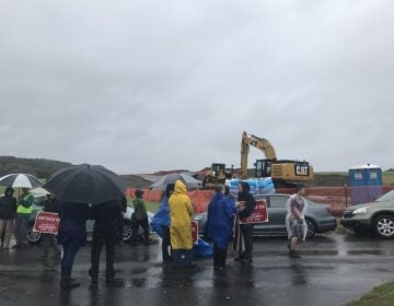 People stand in the rain, behind them a line of cars, behind that, heavy equipment