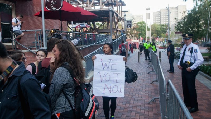 People gather for a protest, a woman holds a sign that says 'we vote no'; to the right a campus police officer