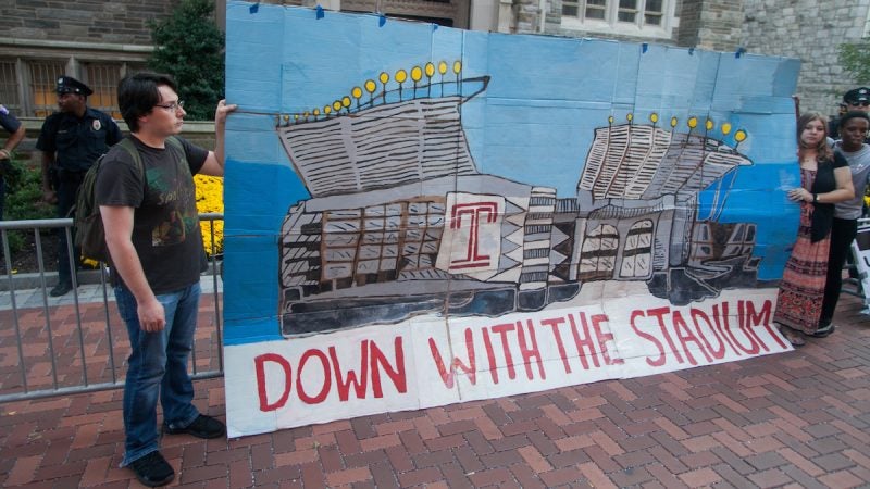 People hold a large, painted cardboard sign depicting the stadium in question, saying 'down with the stadium'