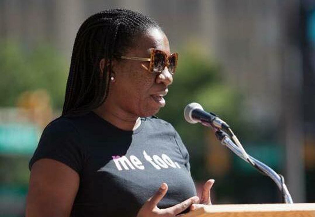 A woman in a black tshirt that reads " me too" speaks at a podium