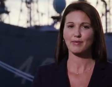 A woman speaks with a warship in the background