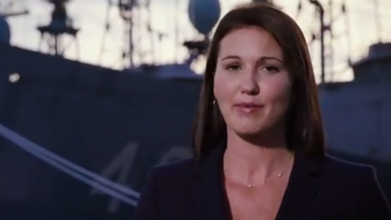 A woman speaks with a warship in the background