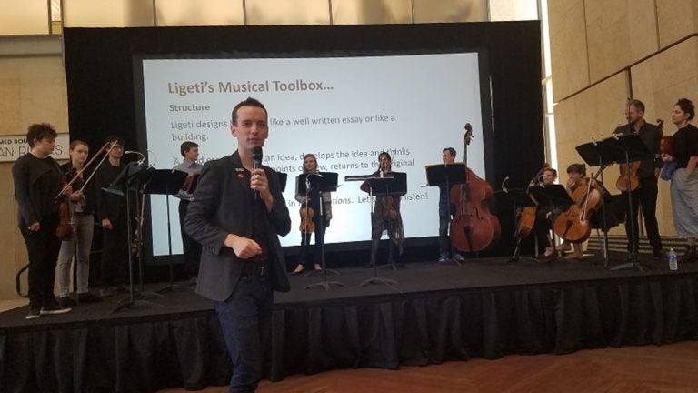 A man with a mic in the foreground, middle-school musicians stand on a stage in the background