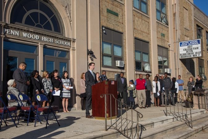 Wide shot of people standing in front of a school, one at a podium