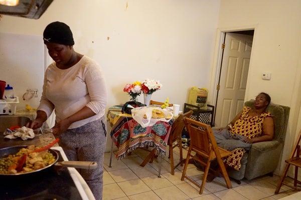 Tatiana Angama, stands at the stove cooking, her mother in the chair off to the right