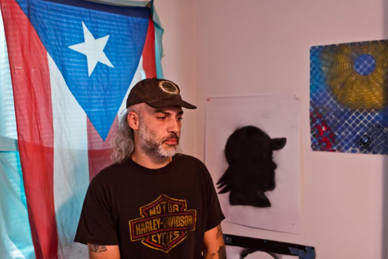A bearded man in a Harley Davidson tshirt and a hat stands in a home; the flag of Puerto Rico hands in the window behind him