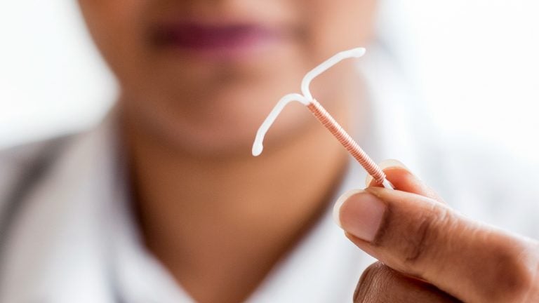 A woman, blurred, and mostly off screen holds an IUD close to the camera (in focus)